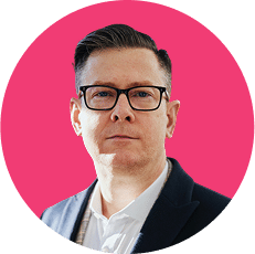 Man with glasses and short hair wearing a dark jacket and white shirt against a pink circular background.